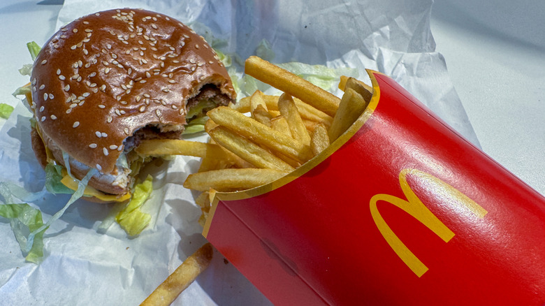A carton of McDonald's french fries laying on a paper wrapper next to a Big Mac with two bites taken out of it