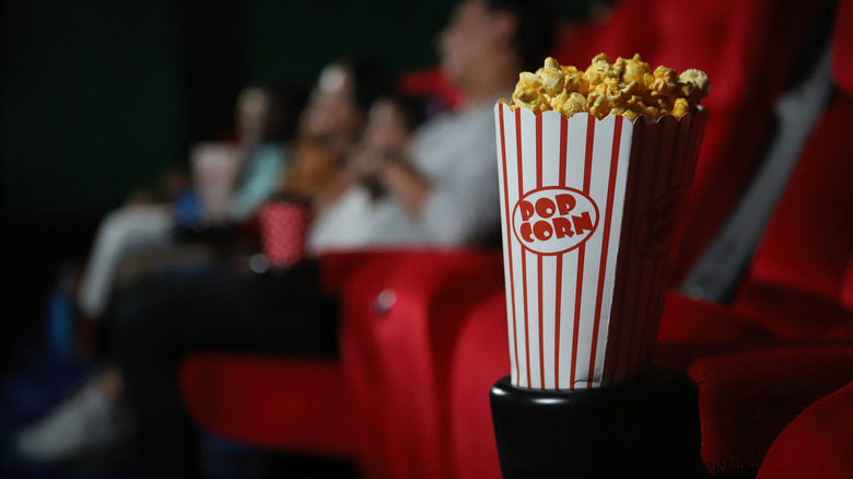 A small bucket of popcorn with moviegoers sitting in read theater chairs in the background