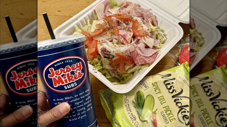 Hand holding Jersey Mike's blue soda cup next to bag of potato chips and "sub in a tub" salad in white takeout container