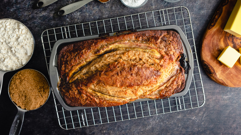 Loaf of banana bread inside pan next to scoops of ingredients like flour and brown sugar