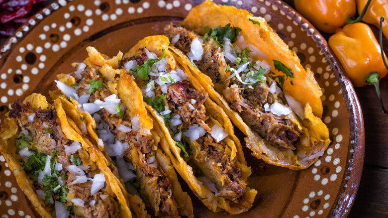 Four hard-shell tacos filled with beef, white onions, and cilantro on a brown ceramic plate.