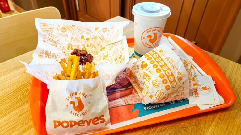 Popeyes food items on orange tray including sleeve of French fries, wrapped sandwich, and drink cup with logo