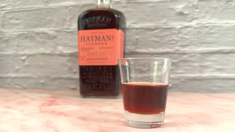 Close-up on shot glass of sloe gin in front of blurred bottle of Hayman's of London on pink table