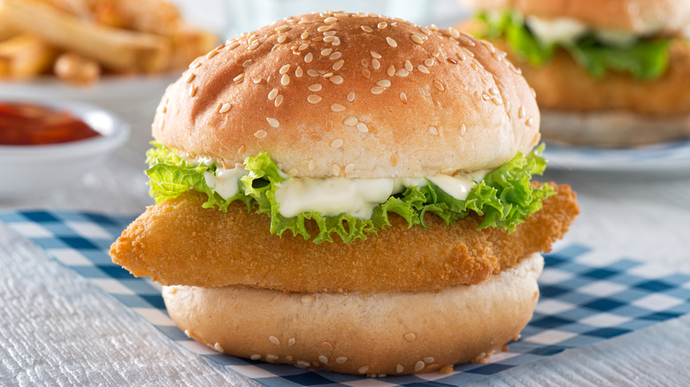 Sandwich with fried fish patty, lettuce, and tartar sauce on round sesame seed bun on white table with blue checkered cloth