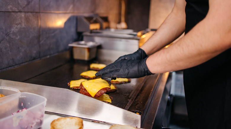 Gloved hands tending to cheeseburgers on a griddle.