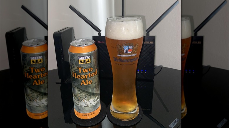 A green and orange tall can of Bell's Two Hearted Ale with an illustration of a trout, next to a tall glass of the amber-colored beer on a glass table.
