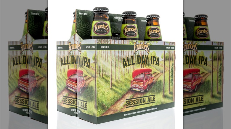 A six-pack containing glass bottles of Founders All-Day IPA on a white background. There's an illustration of a red car, canoe on the roof, driving down a dirt road through the woods, on the packaging.