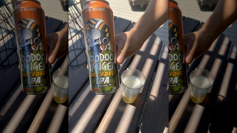 A hand holds a tall, orange can of Voodoo Ranger Juice Force in front of a picnic table. On the table is a glass half full of a heady, golden-colored beer.