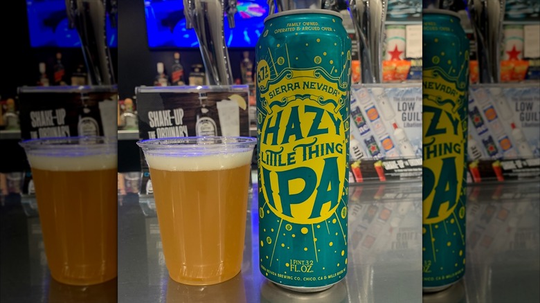 A tall, green can of Hazy Little Thing, next to a plastic cup containing a pour of a golden-colored, cloudy beer.