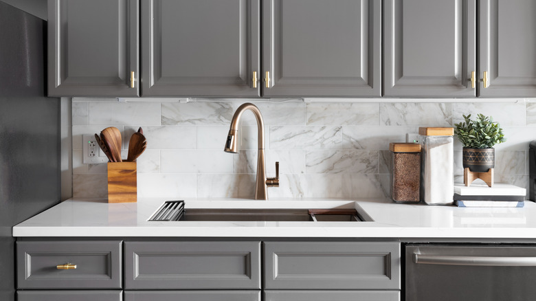 A kitchen sink detail shot with gray cabinets, a white marble countertop and backsplash, and decorations