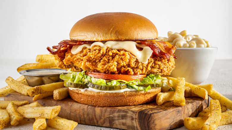 Crispy chicken sandwich with bacon, cheese, tomato, lettuce, and pickles, served on wooden board with fries