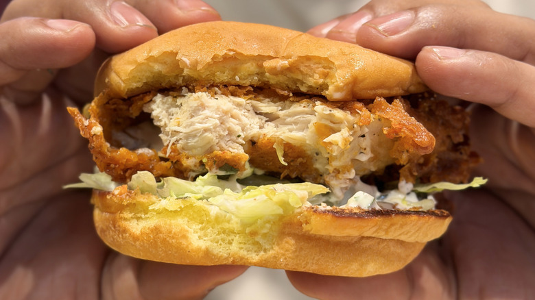 Hands holding crispy chicken, lettuce, and mayo sandwich with bite taken out