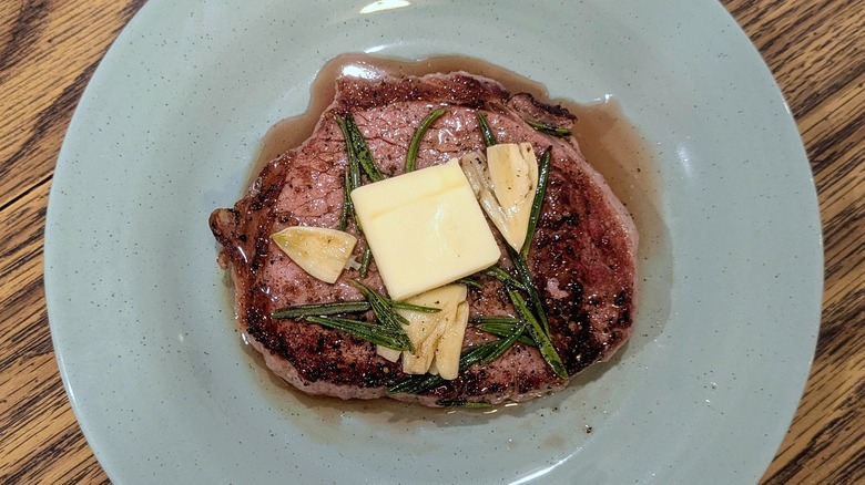 An eye of round steak on a pale blue plate with rosemary, garlic cloves, and a pat of butter