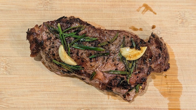 A whole strip steak on a cutting board with rosemary and garlic cloves