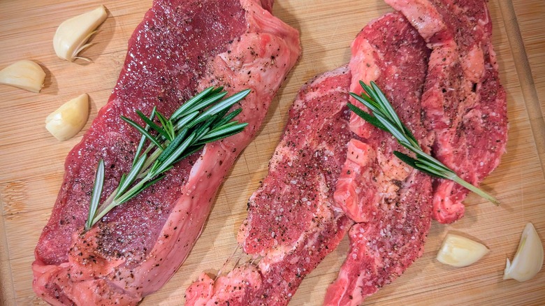 Seasoned raw steaks with sprigs of rosemary and garlic cloves on a cutting board
