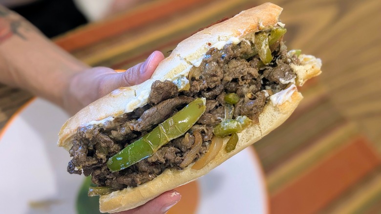 A hand holds a cheesesteak sandwich with onions and green peppers