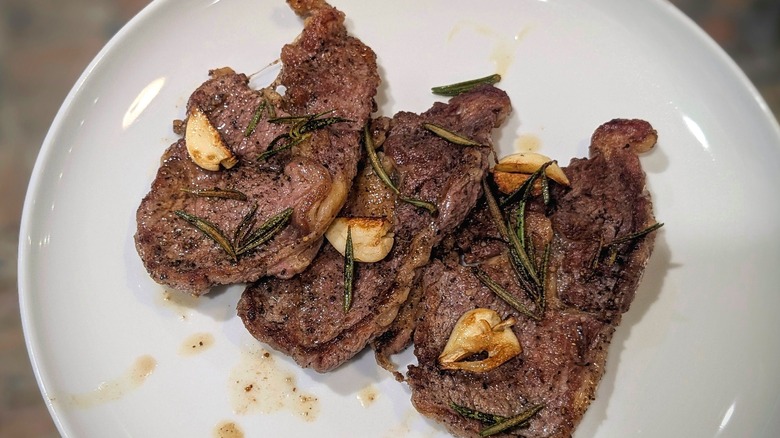 Three thin-cut ribeye steaks with rosemary and garlic cloves on a white plate