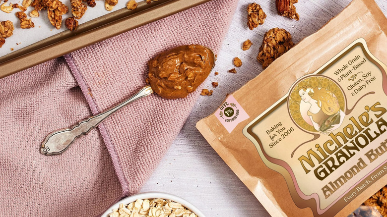 Bag of Michele's Almond Butter Granola alongside spoonful of almond butter, oats, towel, and granola chunks on table