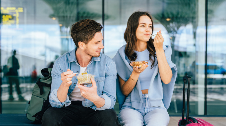 Two young adult friends eating snacks while sitting on the bench at airport terminal
