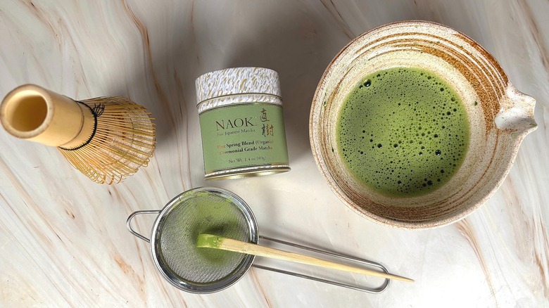 Matcha in chawan next to strainer and bamboo scoop, chasen on holder, and Naoki matcha tin on marble surface
