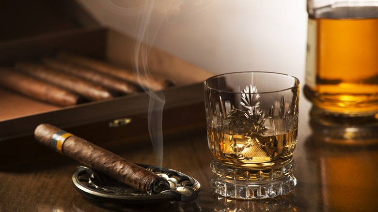 A glass of whiskey next to a lit cigar, with a cigar box and a bottle of whiskey shown in the background