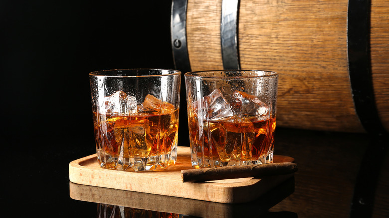 Two filled whiskey glasses on a wooden tray with a cigar placed in front and a barrel shown behind