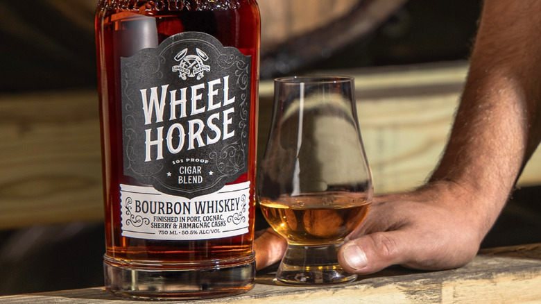 A close shot of a bottle of Wheel Horse Cigar Blend Bourbon with a person holding a filled glass next to it