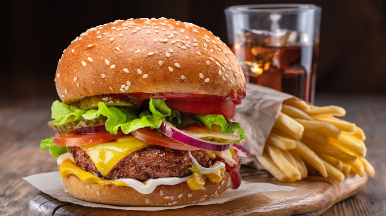 Cheeseburger with vegetables on wooden board with sleeve of french fries and glass of brown soda in background