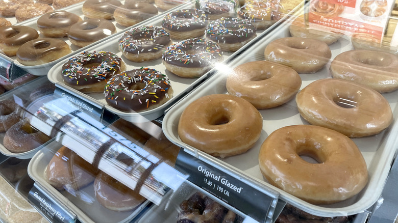 A glass display case at a Krispy Kreme showing trays of donuts including original glazed, chocolate ice glazed, and more