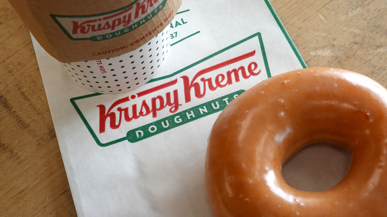 A Krispy Kreme original glazed donut sitting on the edge of a Krispy Kreme doughnits napkin with a coffee in the top left corner