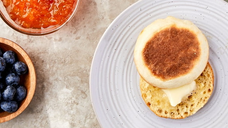 A buttered English muffin sits on plate with fruit and jam nearby