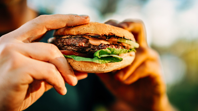 Hands holding a hamburger with cheese, lettuce, and tomatoes