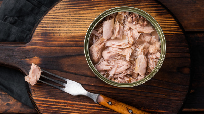 an open can of tuna on a wooden cutting board with a fork beside it skewering a small piece of fish