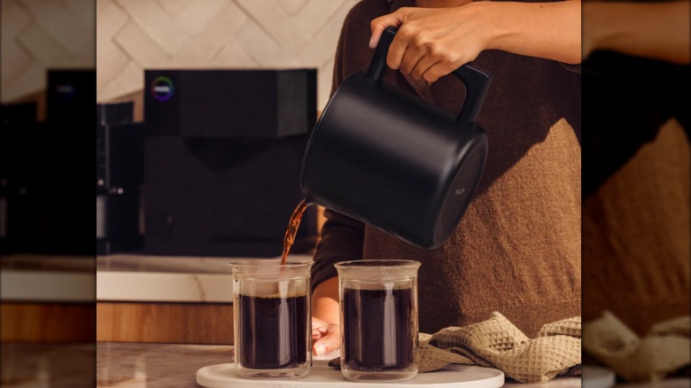 A woman pours two large glasses of coffee from the black carafe of an Aiden Precision brewer