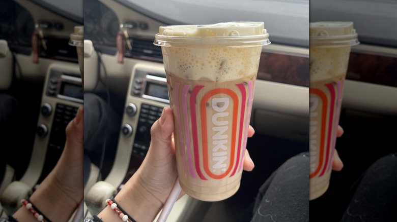 Mirrored image of hand holding Dunkin' banana coffee in front of car