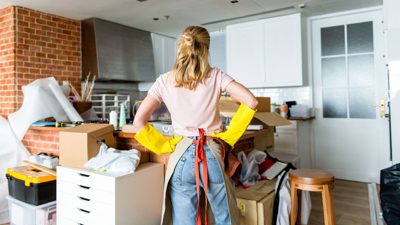 A woman in yellow cleaning gloves looks at a cluttered kitchen