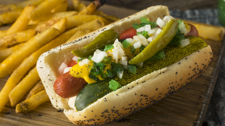 Chicago style hot dog featuring a poppy seed bun, a pickle, yellow mustard, green relish, and onion on a board with some french fries
