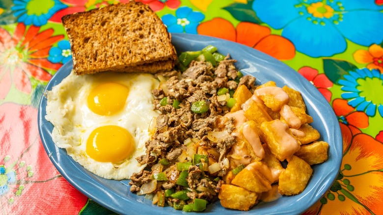 Toast on plate with eggs, potatoes, thin-sliced beef and veg with floral tablecoth background
