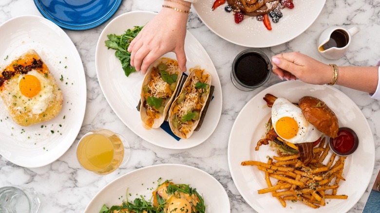 Hands reaching for an assotrment of brunch dishes including tacos, fries, and breakfast sandwich