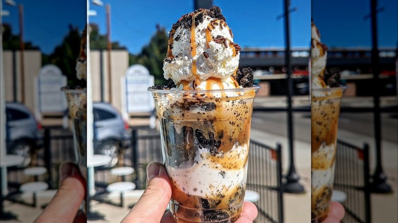 Hand holding an Oreo ice cream sundae with caramel sauce, crumbled Oreos, and sprinkles in a plastic cup