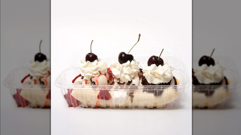 Banana split in a plastic take-out container, topped with whipped cream, cherries, strawberries, and chocolate sauce