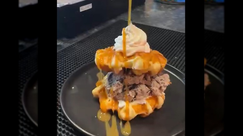 Ice cream sandwiched between two waffles and topped with whipped cream, being drizzled with syrup