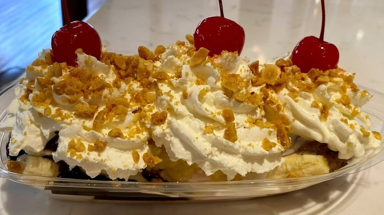 Banana split topped with whipped cream, nuts, and three cherries in plastic takeout container