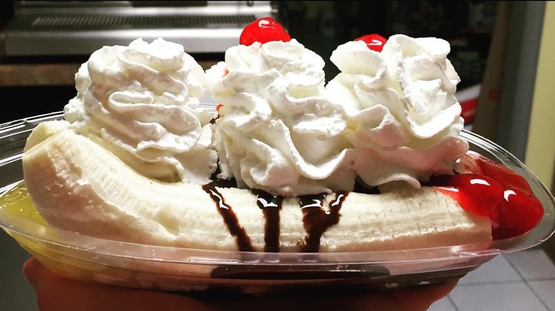 Banana split with whipped cream, chocolate syrup, and cherries in plastic takeout container