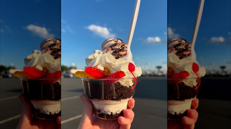 Hand holding sundae with gummy worms and cookie crumbles in clear cup with spoon