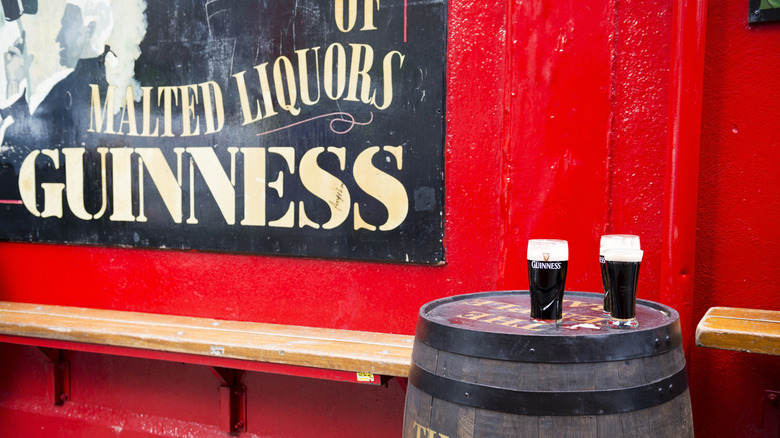 Two pints of Guinness on a barrel in front of red wall and Guinness advertising sign