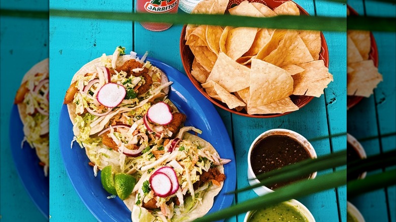 Plate of tacos and bowl of tortilla chips with salsas on bright blue table