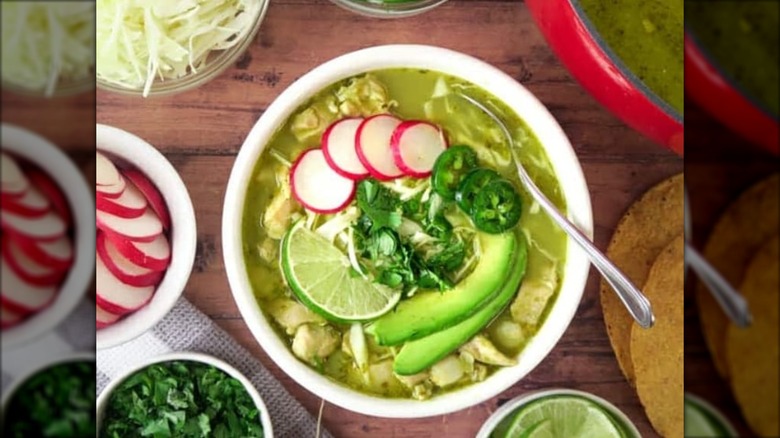 Dish of chicken pozole verde with avocado slices and side dishes of ingredients