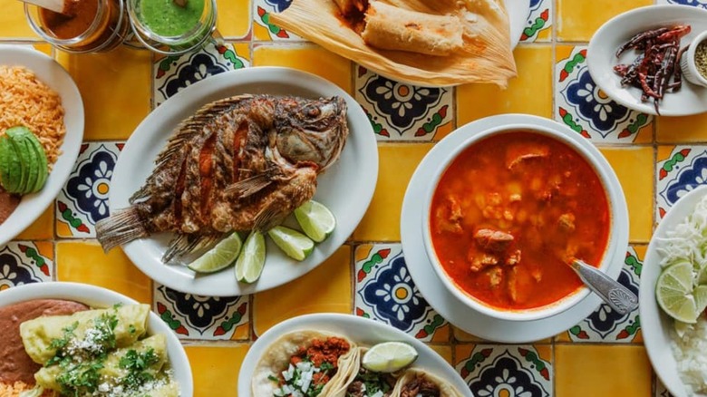 Assorted Mexican dishes including whole fish, soup, tamales, and street tacos on colorful background