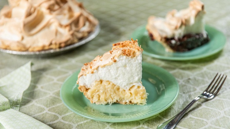 A slice of coconut cream pie with a whole pie and a different pie slice out of focus in the background